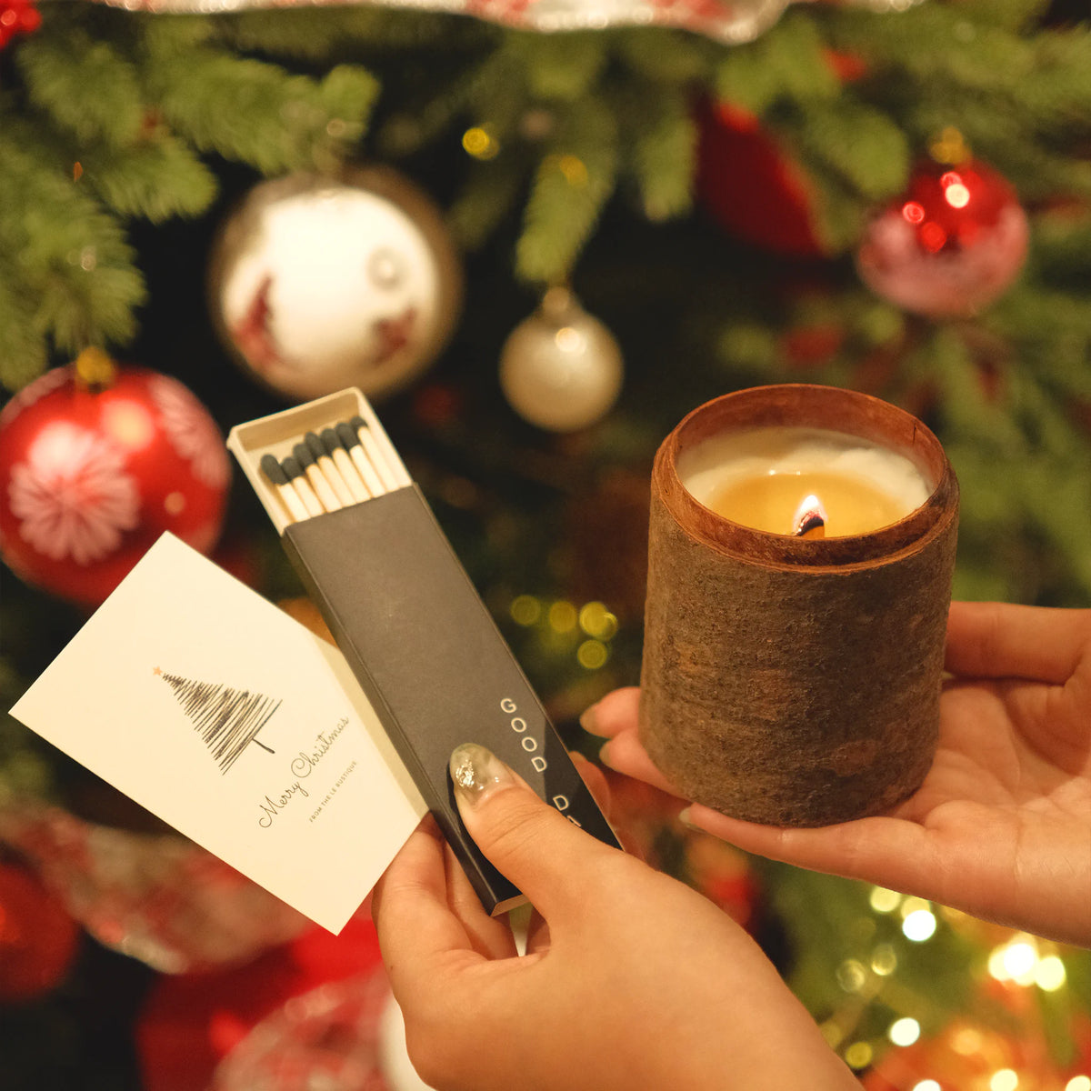 Person holding a essential oil candle and matches in front of a decorated Christmas tree.